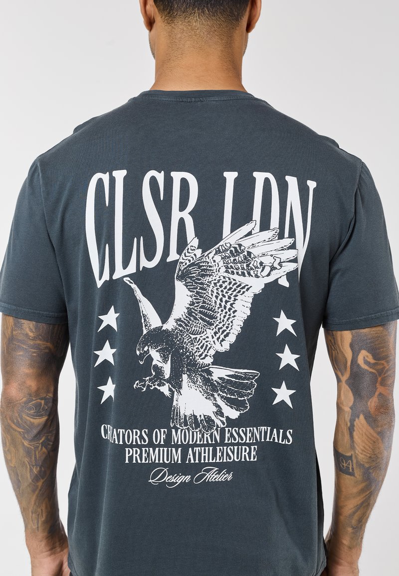 Muškarac s tetovažama nosi tamnosivu majicu s bijelom grafikom orla, zvijezdama i natpisima "CLSR LDN" i "Creators of Modern Essentials."