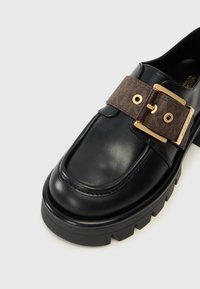 Juodas odinis loaferis su masyviu padu, dekoruotas rudą juostą su auksiniu užsegimu ir logotipo akcentais. Lygus paviršius ir elegantiškas dizainas.