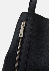 Sac en cuir noir avec une surface texturée, fermeture zippée dorée et longue anse. La tirette de la fermeture éclair a un accent triangulaire.