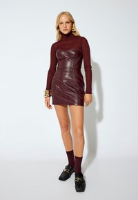 Burgundy turtleneck paita yhdistettynä muotoiltuun burgundinväriseen nahkamaiseen mini-mekkoon, jossa on sydänkaarrekaula-aukko ja sileä tekstuuri. Kultaostekorut ja mustat loafersit viimeistelevät asun.