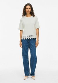 Top gris claro de manga corta con un dobladillo de encaje, combinado con jeans azules y zapatos beige. La tela parece suave con un ajuste relajado.