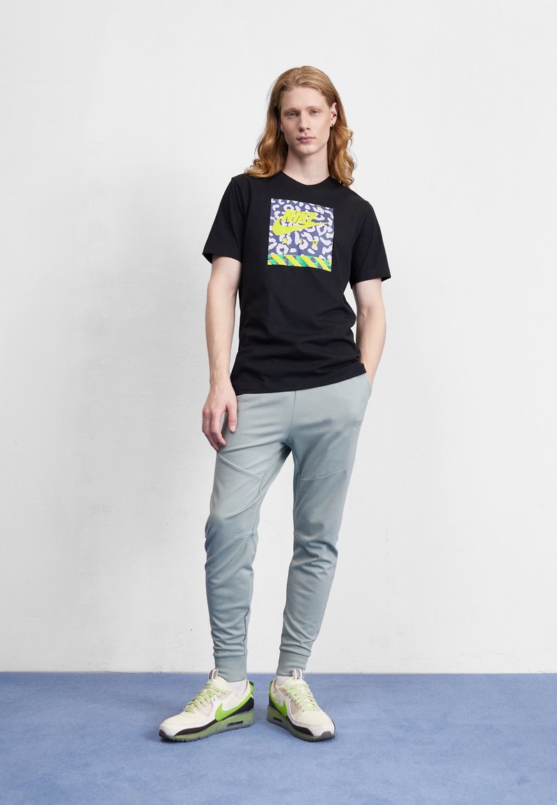 T-shirt noire avec impression graphique colorée, joggings gris clair, et baskets blanches avec accents verts. Texture lisse, coupe décontractée.