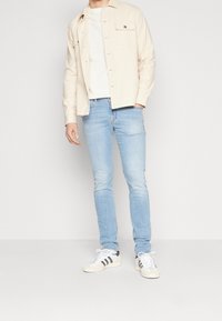 Ljusbeige knapp-up skjorta över en vit t-shirt, tillsammans med ljusblå slim-fit jeans och vita sneakers med svarta ränder.