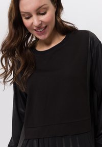 Glunderende vrouw met lang bruin haar, gekleed in een zwart top met lange mouwen met satijnen mouwen en een geplooide onderzoom tegen een witte achtergrond.