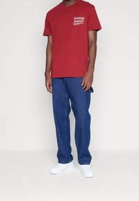 T-shirt en coton rouge avec un logo blanc sur la poitrine, associé à un jean en denim bleu. La tenue est complétée par des baskets blanches.