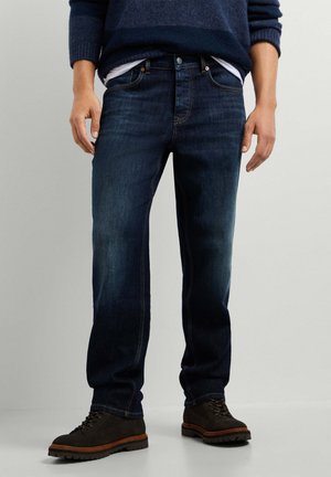 Homme portant un jean bleu foncé, un pull bleu marine, un sous-vêtement blanc et des bottes lacées marron foncé, debout devant un fond uni.
