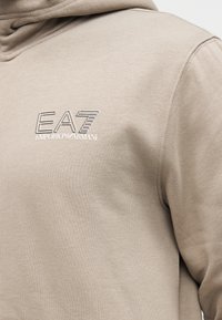 Beige hoodie van zachte stof met een geborduurd logo "EA7 EMPORIO ARMANI" aan de voorkant in een contrasterende kleur. Eenvoudig ontwerp met een klassieke pasvorm.