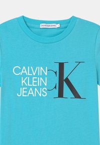 Turkusowa koszulka z bawełny z napisem "CALVIN KLEIN JEANS" w białym kolorze oraz dużym czarnym logo "K" na przedzie. Okrągły dekolt.