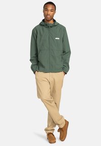 Grüne, leichte Jacke mit Kapuze, Reißverschluss vorne und elastischen Bündchen, kombiniert mit beigen Hosen und braunen Wildlederschuhen.