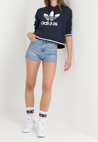 Námornícký zkrácený svetr s bílým logem Adidas, roztrhané džíny šortky a vysoké ponožky po kolena s růžovými a námornickými pruhy a hvězdami. Bílé tenisky.