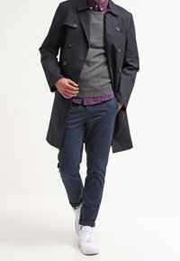Marineblauer Trenchcoat mit doppelreihigen Knöpfen, grauem Pullover, karierter Hemd und dunkeln Hosen. Weiße Sneaker runden den Look ab.