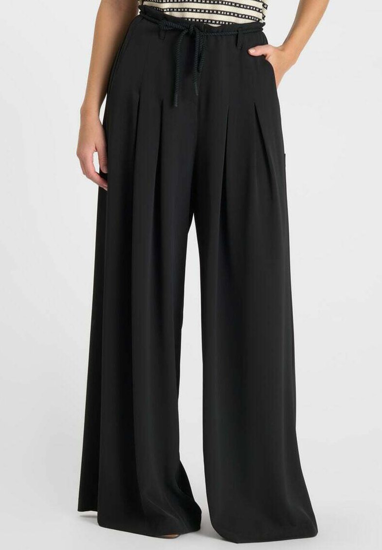 Femme portant un pantalon large noir taille haute avec plis avant et cordon de serrage, main reposant dans la poche droite, haut court noir et blanc.