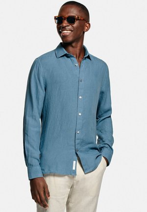 Lachende man met schildpaddenzonnebril, blauwe blouse met lange mouwen en lichte beige broek, met één hand in de zak.