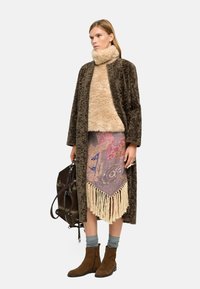 Długi, brązowy, teksturowany płaszcz na beżowym futrzanym golfie, wzorzysta fioletowa spódnica z frędzlami, szare skarpetki i brązowe botki na kostkę. Brązowa torba.