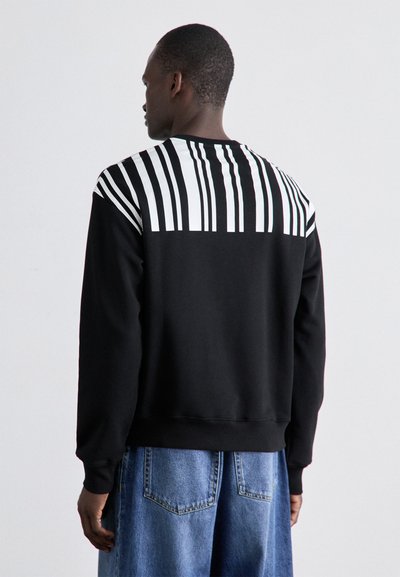 Sweatshirt preto com um padrão de riscas brancas na parte superior das costas e ombros. Apresenta mangas longas e um corte descontraído. Calções de ganga visíveis por baixo.