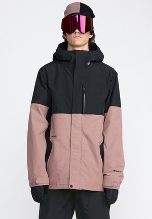 Veste imperméable noire et rose avec capuche, poche zippée et étiquette GORE-TEX. Portée avec un bonnet coloré et des lunettes de soleil miroir.