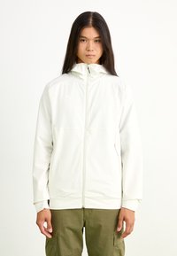 Chaqueta ligera de color crema con cierre de cremallera frontal, cuello alto, puños elásticos y dos bolsillos laterales. Presenta un sutil detalle de logo.