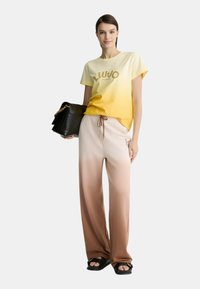 T-shirt ombre giallo-arancio con logo dorato, abbinato a pantaloni ombre beige-marroni. Una borsa nera e sandali completano l'outfit.