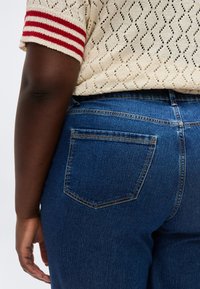 Κοντινό πλάνο ενός ατόμου που φοράει σκούρο μπλε τζιν παντελόνι και μπεζ πλεκτό πουλόβερ με κόκκινες ρίγες στο μανίκι.