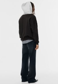 Felpa nera oversize con cappuccio grigio chiaro, indossata sopra una camicia a quadri, abbinata a jeans blu scuro a taglio ampio.