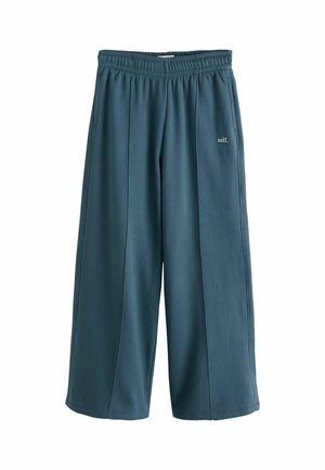 Weit geschnittene dunkel petrolefarbene Jogginghose mit elastischem Bund und dem Text "self." am oberen linken Oberschenkel.