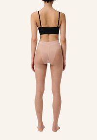Schwarzer Bralette und durchsichtige, hautfarbene, hochgeschnittene Strumpfhosen mit Netzpaneelen und glatter Textur, von hinten vor einem beigen Hintergrund betrachtet.