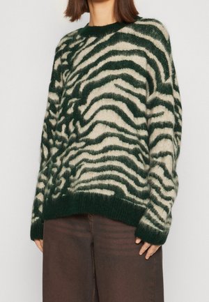 Femme portant un pull doux rayé zèbre vert foncé et crème à manches longues, avec un pantalon marron foncé, debout devant un fond uni.