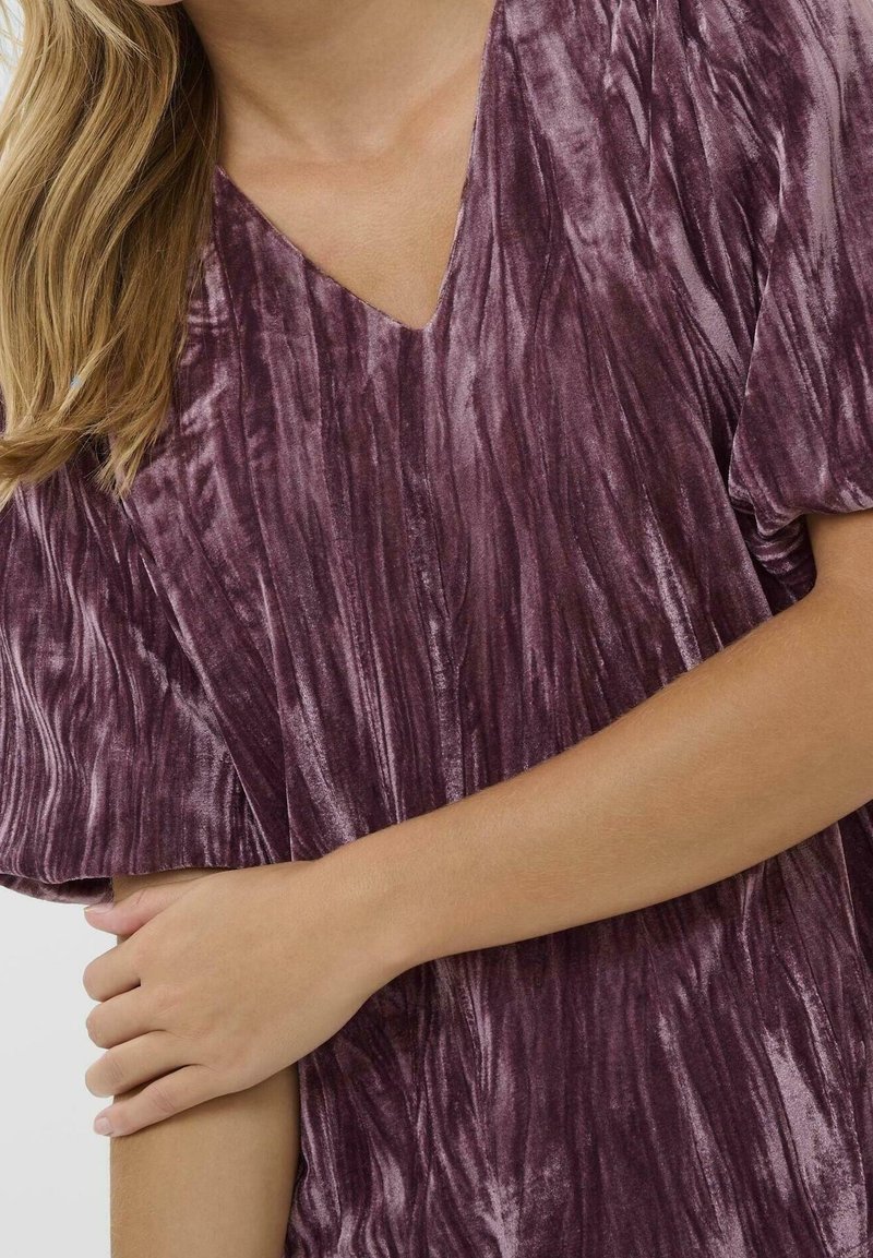 Femme aux longs cheveux blonds porte un haut en velours violet froissé à col en V, croisant un bras sur son corps devant un fond uni et clair.