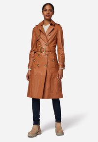 Brown leather trench coat with a belted waist, double-breasted buttons, and shoulder epaulets, paired with dark jeans and beige ankle boots.