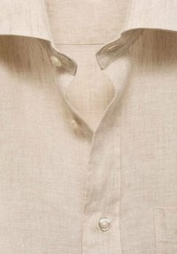 Zbliżenie na kołnierzyk beżowej lnianej koszuli i przednie guziki, podkreślające teksturę tkaniny oraz detale guzików.