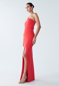 Punainen, olkaimeton mekko, jossa on istuva malli ja korkea halkio. Mekossa on sileä rakenne ja jäsennelty siluetti. Asustettu kultaisilla remmikkäillä koroilla.