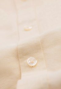 Gros plan sur un tissu de chemise beige clair montrant un petit bouton nacré blanc cousu avec un fil assorti.
