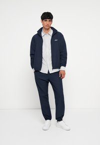 Navy blue lightweight jacket with a high collar, zipped front, and elastic cuffs, paired with light gray striped shirt and dark pants. White sneakers.