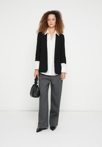 Μαύρο σακάκι ραμμένο στα μέτρα πάνω από λευκό πουκάμισο, γκρι παντελόνι με φαρδιά μπατζάκια και μαύρα παπούτσια με μυτερή μύτη. Μαύρη τσάντα με υφή στην επιφάνεια.