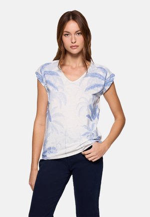 Mujer de pie con la mano en el bolsillo de unos pantalones azul marino, vistiendo una blusa blanca de manga corta con estampado de hojas de palma azules.