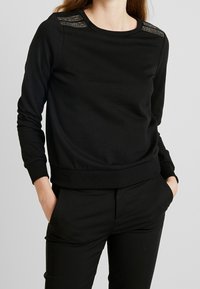 Sweat-shirt noir en tissu doux, avec un col rond, des manches longues et des accents texturés sur les épaules.