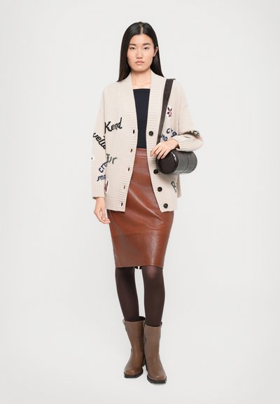 Cardigan beige con motivi testuali, bottoni neri, abbinato a una gonna a matita in finta pelle marrone, collant scuri e stivali marroni fino al polpaccio. Borsa in tinta.