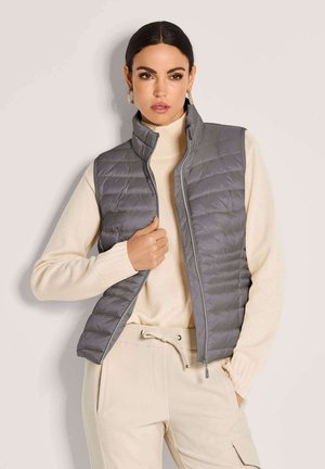 Frau trägt graue gesteppte Weste über cremefarbenem Rollkragenpullover und beigen Hosen mit Kordelzug, steht vor schlichtem hellem Hintergrund.