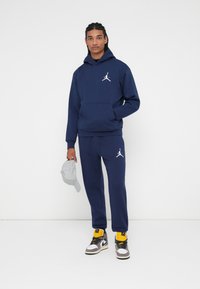 Hanorac din bumbac bleumarin și pantaloni de trening asortați, cu accente albe ale logo-ului. Modelul ține o șapcă gri deschis; poartă adidași negri cu galben.