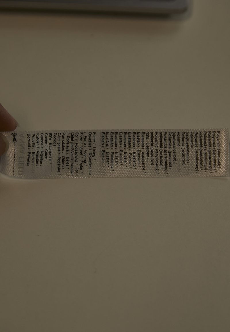 Tøjlabel med angivelse af stofsammensætning på flere sprog, herunder procentdele af bomuld, polyester og elastan, holdt op mod en lys baggrund.