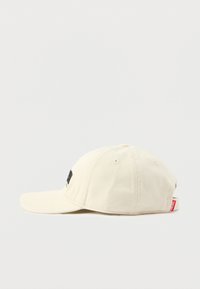 Μπεζ βαμβακερό καπέλο με καμπυλωτή γείσο, μαύρη κεντημένη λογότυπο στο μπροστινό μέρος και κόκκινη ετικέτα στο πίσω μέρος. Υφασμάτινη υφή και δομημένος σχεδιασμός.