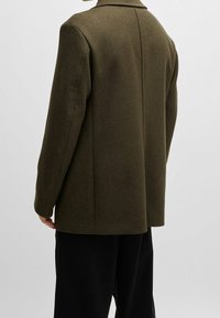 Olivgrön ullblandad blazer med strukturerad design, slagkrage och två bakventiler, i kombination med svarta byxor. Texturerad tygfinish.