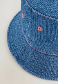 Chapeau seau en denim bleu avec des coutures contrastantes roses et des accents de boutons, présentant un tissu texturé et une large visière arrondie.