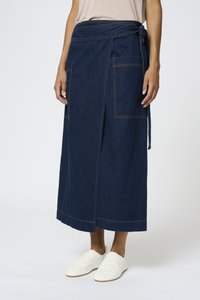 Denim omotana suknja u tamnoplavoj boji s dva velika prednja džepa i detaljem vezivanja u struku. Uparena s bijelim tenisicama za ležeran izgled.