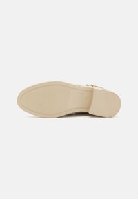 Semelle de chaussure beige clair, texturée avec des nervures et un bord lisse. Présente un petit talon carré et un logo de marque embossé près de la zone des orteils.