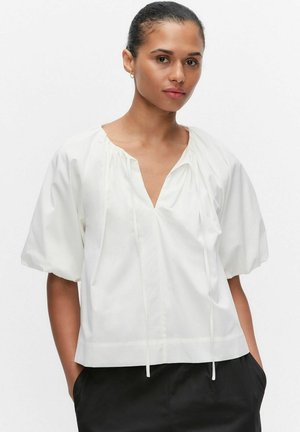 Nainen, jolla on tummat hiukset nutturalla, pukeutunut löysään valkoiseen puseroon, jossa on pöyheät lyhyet hihat, ja mustiin housuihin, seisoo kädet taskuissa.