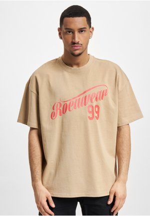 Noormees lühikeste juustega, kandes beežikat üle suuruses T-särki punase tekstiga "Rocnwear 99", seisab tavalise valge tausta ees.