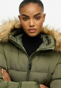 Chaqueta acolchada verde oliva con capucha forrada de piel sintética, cierre de cremallera y textura acolchada. Cuello alto negro debajo y brazos cruzados.