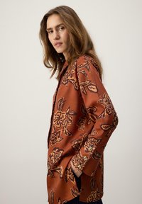 Bluza maro vaporoasă cu model floral negru, detalii cu nasturi la manșete, mâneci lungi și croială lejeră, având o textură netedă.