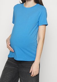 Modré tričko s krátkým rukávem z měkké tkaniny, s kulatým výstřihem a volným střihem, nošené přes těhotenské bříško. Viditelné šedé kalhoty.